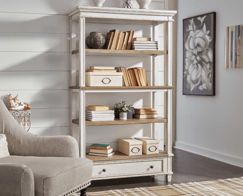 Alt text: "A rustic white bookshelf filled with neatly arranged books, storage boxes, and small decorative items, complementing the elegant decor of a living room with a gray tufted armchair and floral wall art.