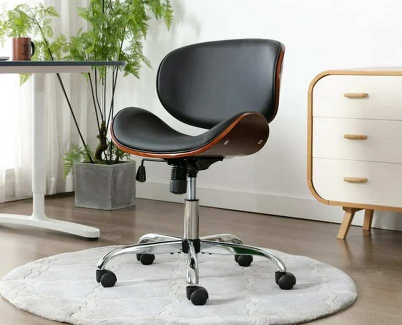 Alt text: "A stylish mid-century modern office chair with a black seat, wooden back, and chrome base, displayed on a circular gray area rug in a bright room with green plants and a white-and-wooden drawer unit.