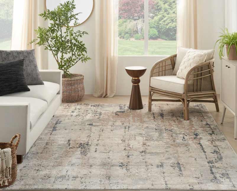 A serene living room corner with natural light filtering through sheer curtains, featuring a comfortable white sofa with gray cushions, a stylish cane armchair with a throw pillow, a decorative bronze side table, and a large distressed rug in muted tones, all contributing to a peaceful and elegant ambiance.