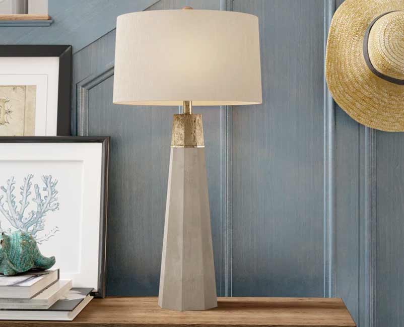 A stylish interior scene with a contemporary table lamp featuring a tapered base and a gold accent, set on a wooden console table against a textured blue wall, accompanied by a woven sun hat and framed artwork, evoking a chic coastal aesthetic.